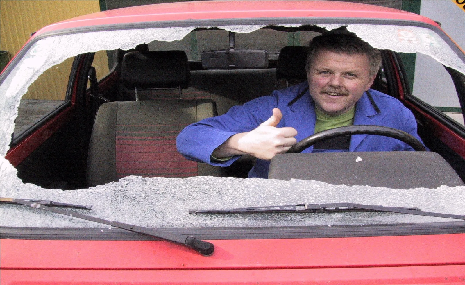 Mann im blauen Arbeitsmantel sitzt hinter einer herausgeschlagenen Windschutzscheibe eines roten Autos und zeigt den Daumen nach oben